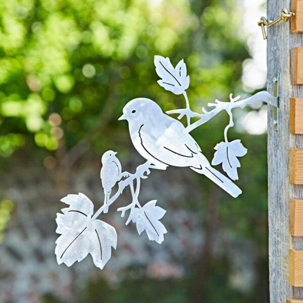 The Recycled Iron Bird Silhouette Garden Art features two birds on a leafy branch, attached to a wooden post, with blurred greenery in the background.
