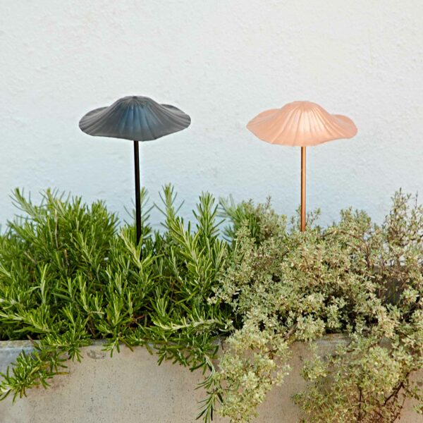 Two Recycled Metal Mushroom Garden Stakes—one black and one pink—add a charming touch to green plants in a concrete planter, standing out against a white wall.