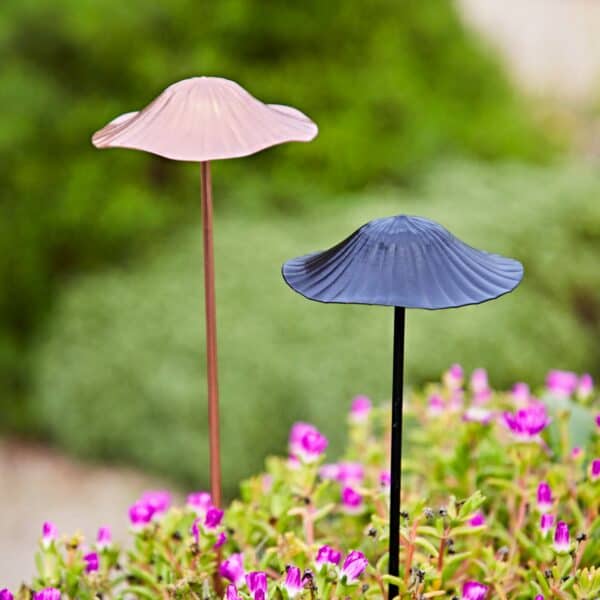 Two Recycled Metal Mushroom Garden Stakes, one pink and one black, stand among lush green foliage and pink flowers, adding whimsical charm to the garden with a softly blurred background.