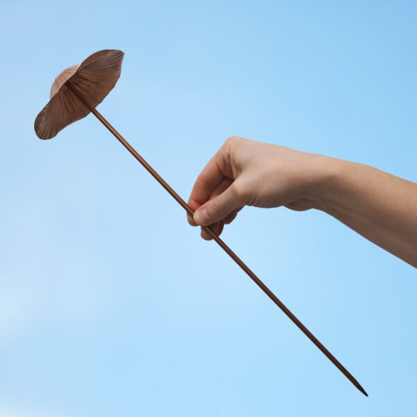 A hand holds a Recycled Metal Mushroom Garden Stake, featuring a long, thin design with a copper mushroom top—perfect eco-friendly decor for your garden, shown against a clear blue sky.