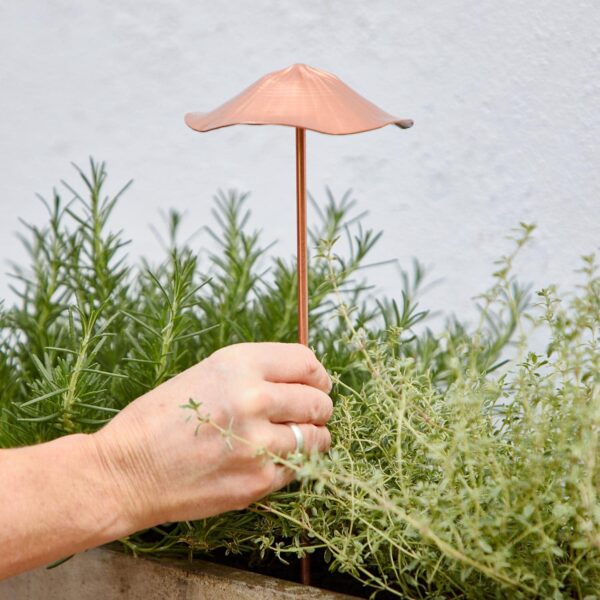 A hand places a Metal Bird Garden Stake Ornament into a bushy green herb garden, adding a whimsical touch to the lush landscape.