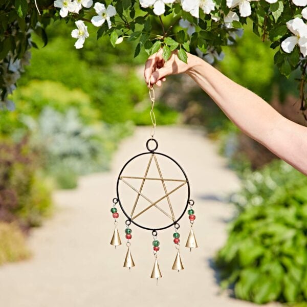 A person holds the Recycled Iron Star and Bells Wind Chime, featuring bells and beads, while standing on a garden path beneath blooming branches.