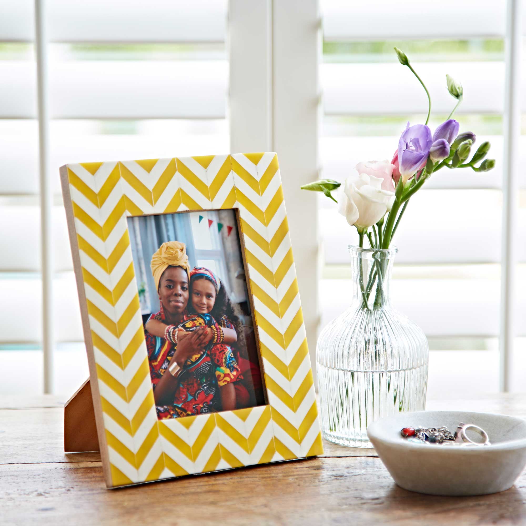 A framed photo with a yellow chevron pattern, perfect as a Mother's Day gift, sits alongside a vase of flowers and a small dish on a wooden table, with window shutters in the background.