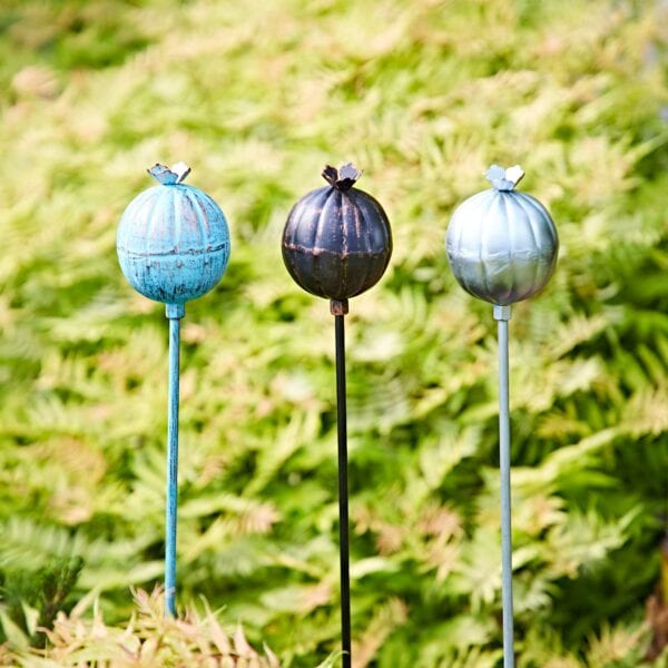 Three Recycled Iron Poppy Seed Head Garden Stake Decorations stand among green foliage, each ornament crafted from recycled iron and shaped like a poppy seed head on a slender stake.