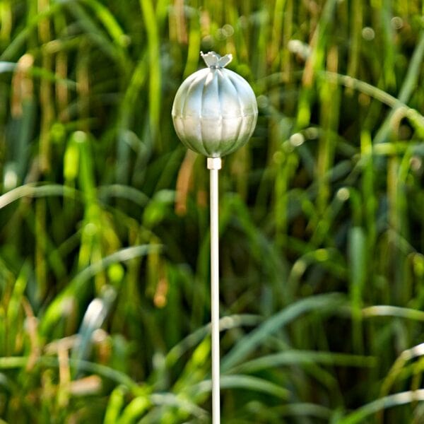 The Recycled Iron Poppy Seed Head Garden Stake Decoration stands gracefully in front of tall green grass.