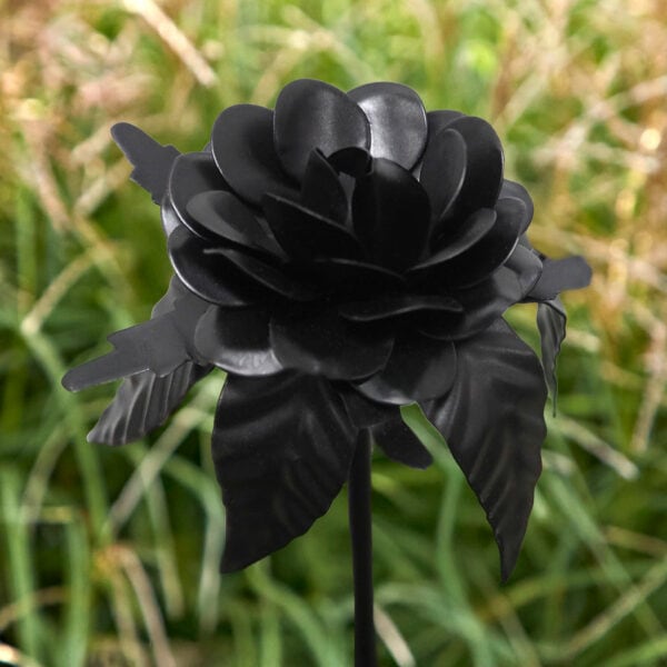 The Black Rose Metal Garden Stick features layered metal petals and leaves, standing outdoors as bold garden decor against a backdrop of green grass and plants.