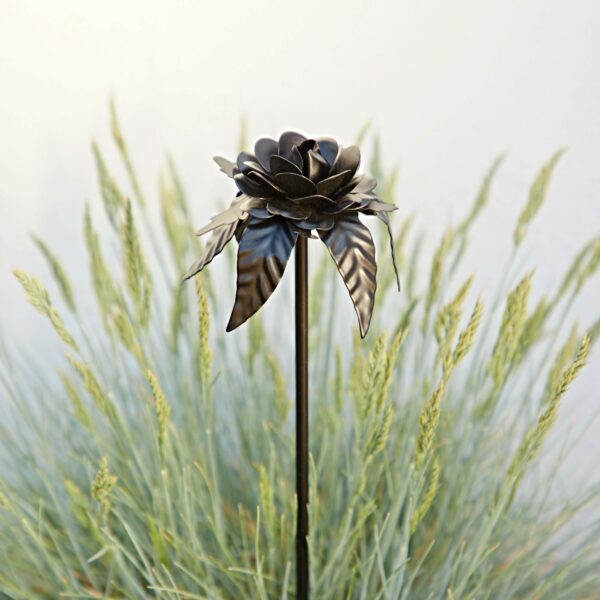 The Recycled Iron Black Rose Garden Stake features a slim stem and bold design, creating striking decor against green ornamental grass with a light, neutral background.