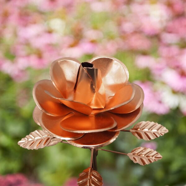 A close-up of the Recycled Metal Copper Rose Garden Stake, featuring metallic leaves and crafted from recycled metal, set against a blurred background of pink and green foliage.
