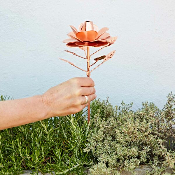 A hand holds the Recycled Metal Copper Rose Garden Stake above green plants, set against a light-colored wall.