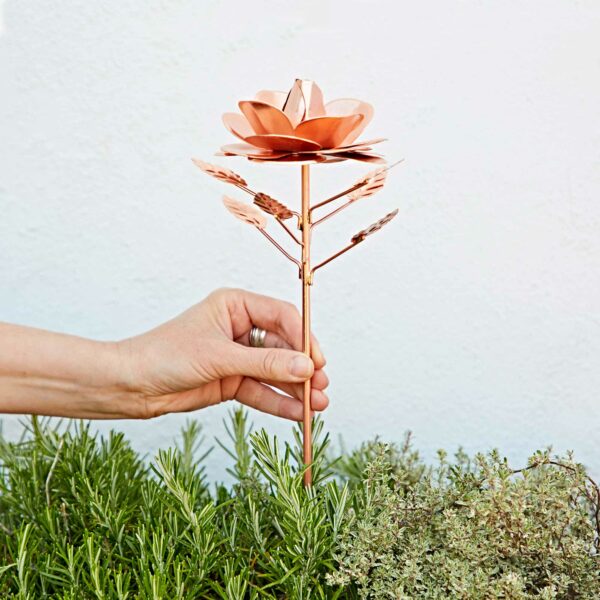 A hand is placing the Recycled Metal Copper Rose Garden Stake, with copper-colored petals, into a lush green garden bed.