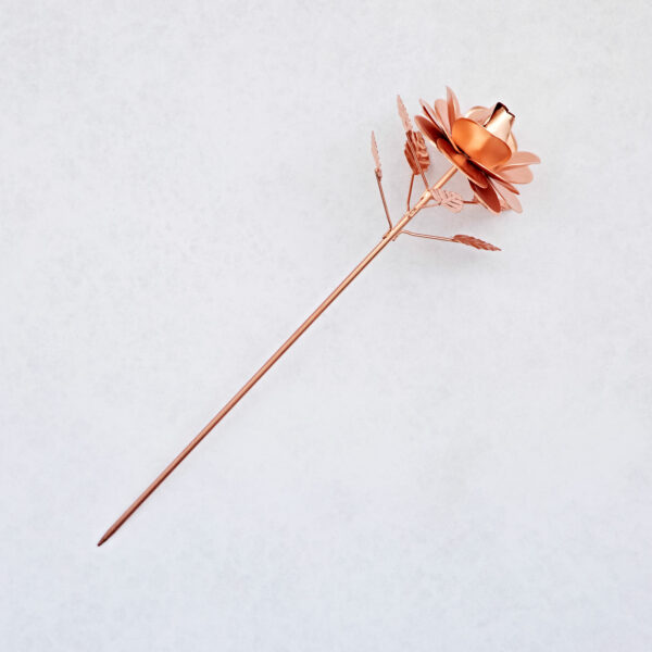 The Recycled Metal Copper Rose Garden Stake features a detailed metallic copper rose with intricate petals and leaves on a long stem, beautifully crafted from recycled metal and shown on a light, textured background.