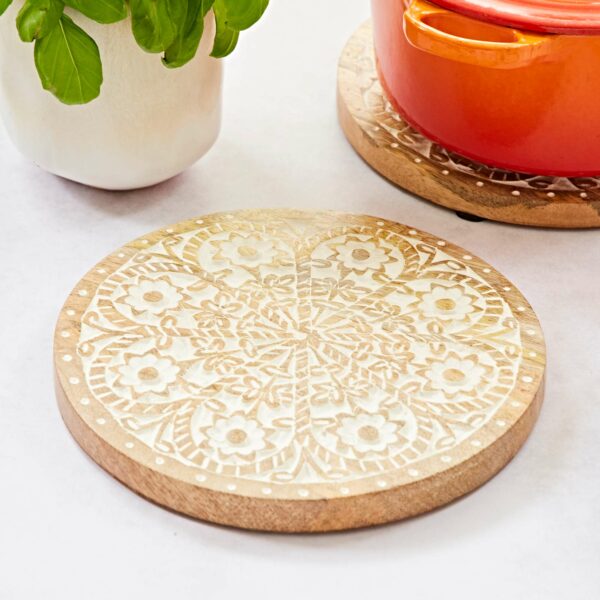 The Round Floral Carved Mango Wood Trivet, featuring an intricate floral design, rests on a white surface near a potted plant and an orange pot.