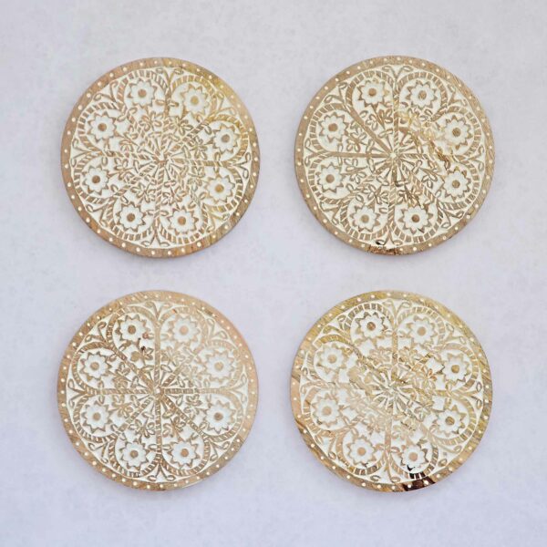 Four round mango wood trivets with intricate floral carvings and geometric cutouts, arranged in a square on a light background. Product: Round Floral Carved Mango Wood Trivet.