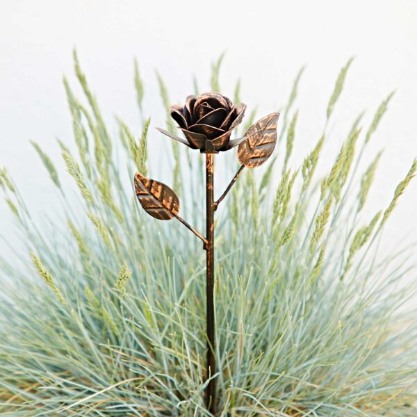 The Recycled Iron Distressed Copper Rose Garden Stake, featuring two leaves, stands in front of tall green grass against a light background, creating a striking accent for your garden.