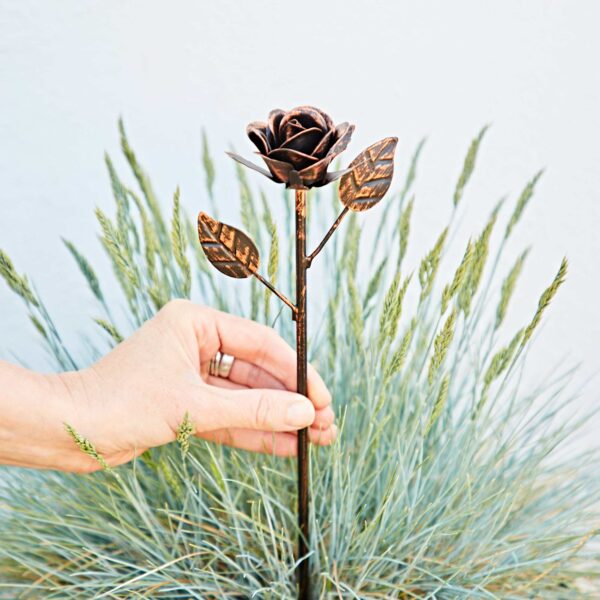 A hand holds the Recycled Iron Distressed Copper Rose Garden Stake with leaves, set against ornamental grass, creating a unique garden accent.