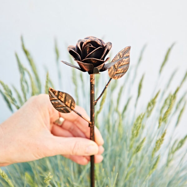 A hand holds the Recycled Iron Distressed Copper Rose Garden Stake, featuring two leaves, against a backdrop of green grass-like plants and a plain light wall.