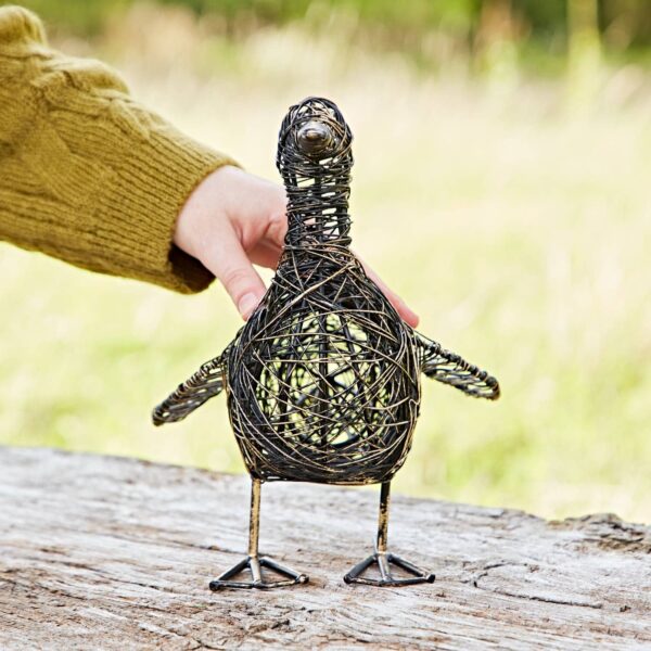 A hand places Anka the Drinking Duck Wire Garden Ornament on a wooden surface outdoors, showcasing its simple and rustic design.