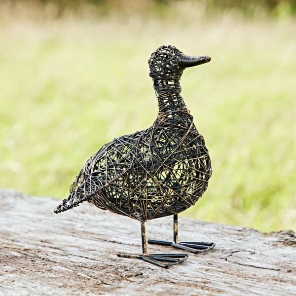 The Anka the Drinking Duck Wire Garden Ornament perches elegantly on a log against a blurred grassy background, offering an exquisite woven metal sculpture for your garden.
