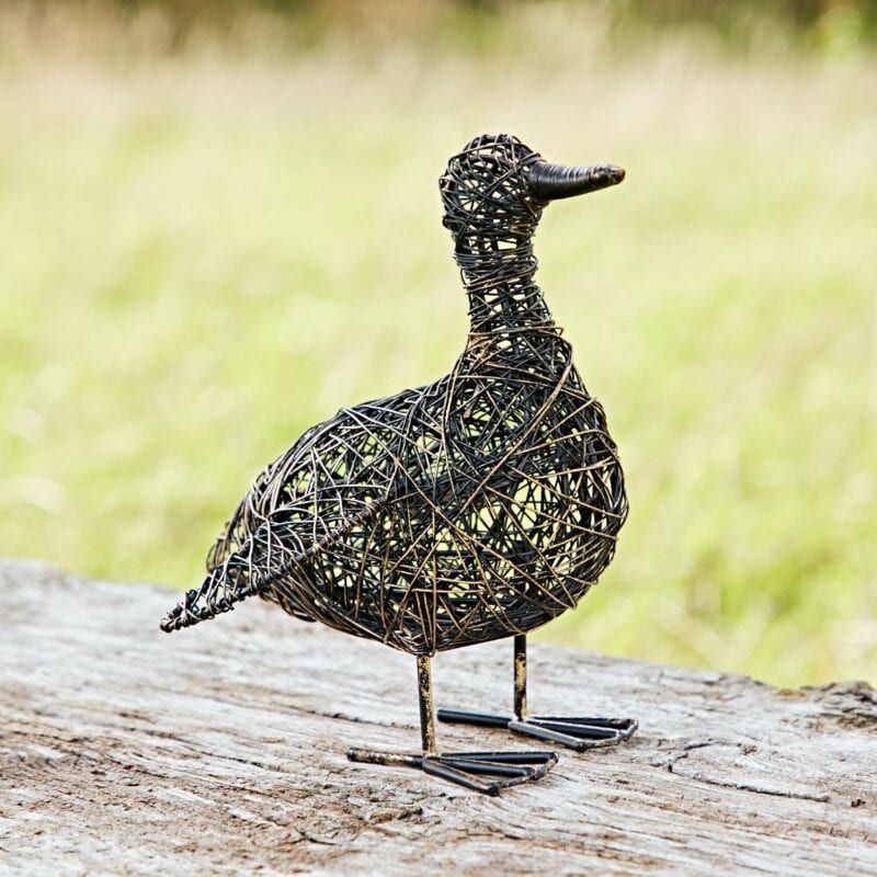 The Anka the Drinking Duck Wire Garden Ornament perches elegantly on a log against a blurred grassy background, offering an exquisite woven metal sculpture for your garden.