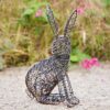 The Hare Wire Garden Ornament, shaped like a rabbit, rests on gravel with green grass and pink flowers in the background.