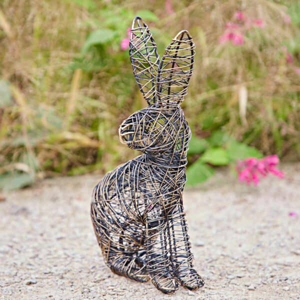 The Hare Wire Garden Ornament, featuring a sitting rabbit, is displayed on gravel with green grass and pink flowers in the background.