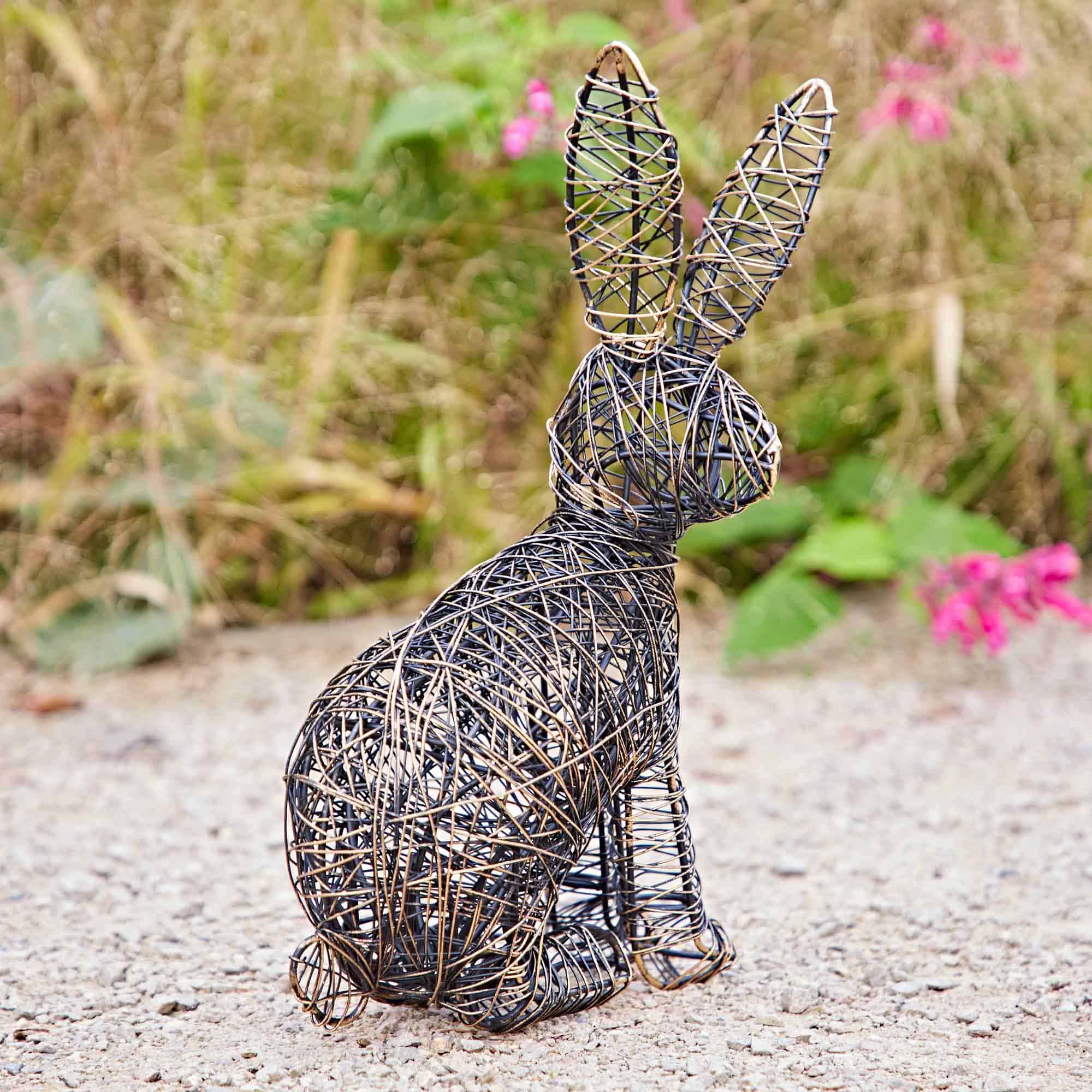 The Hare Wire Garden Ornament, a rabbit sculpture, is placed on gravel with green plants and pink flowers in the background.