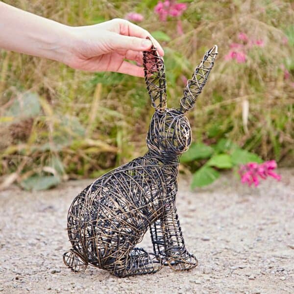 A hand holds the ear of a Hare Wire Garden Ornament, shaped like a rabbit, positioned outdoors on grass with pink flowers in the background.