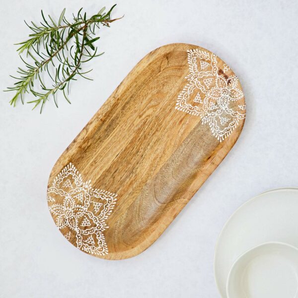 A Round Floral Carved Mango Wood Trivet, adorned with white mandala corner designs, is elegantly placed next to a sprig of rosemary and white plates on a light surface.