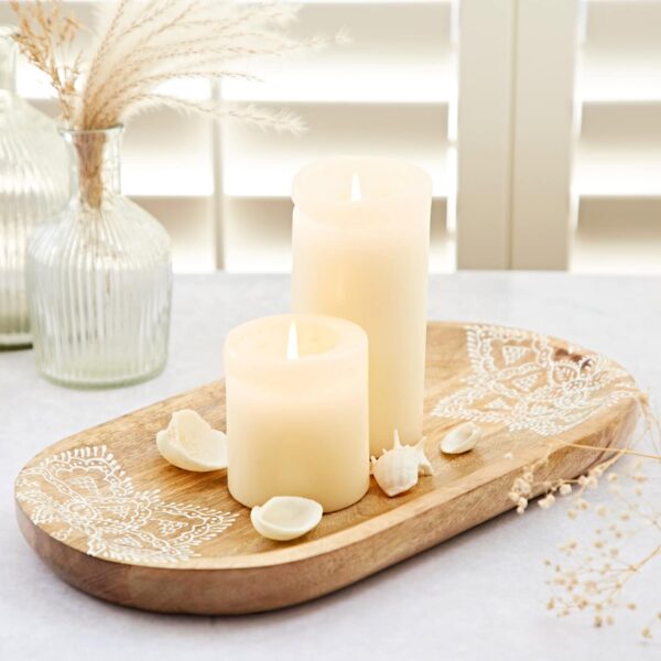 Two lit candles of different sizes sit on a Round Floral Carved Mango Wood Trivet, embellished with decorative shells and patterns, on a light-colored table. The backdrop includes dried plants in vases and window blinds, enhancing the elegance of the scene.