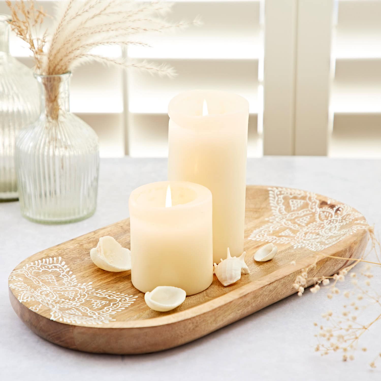 Two lit candles of different sizes sit on a Round Floral Carved Mango Wood Trivet, embellished with decorative shells and patterns, on a light-colored table. The backdrop includes dried plants in vases and window blinds, enhancing the elegance of the scene.