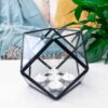 A Triangular Recycled Glass Wall Terrarium crafted from recycled glass holds five glowing tealight candles on a white surface, with a lush plant and textured blanket enhancing the warm backdrop.