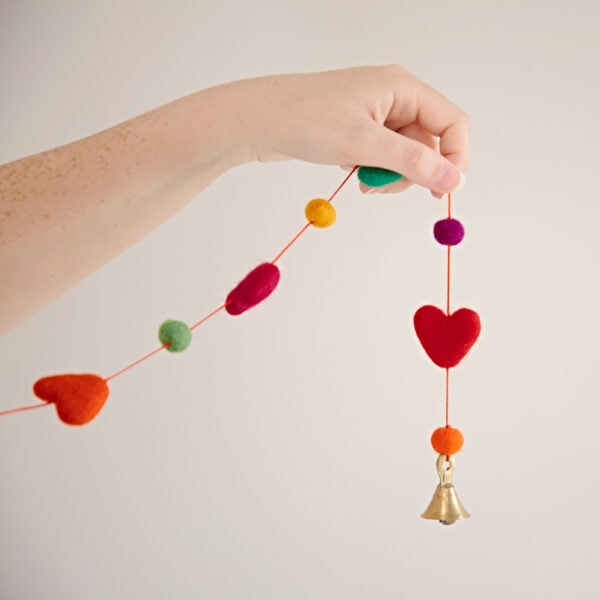A hand holds a "Felt heart Ball Garland," showcasing colorful felt decorations with hearts and round shapes, finishing with a small brass bell.