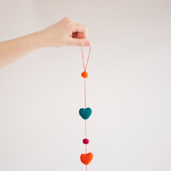 A hand holds a Felt heart Ball Garland featuring orange circle, blue heart, pink circle, orange heart, and orange circle shapes. The vibrant display stands out against the plain white background.