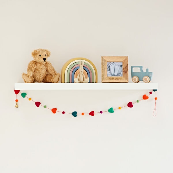 A white shelf showcases a teddy bear, rainbow decor, framed baby feet, and toy truck. Below, a colorful heart garland elegantly hangs.