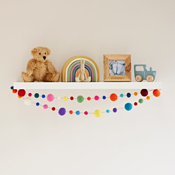 A white shelf showcases a teddy bear, a wooden rainbow, a photo frame, a blue toy vehicle, and Felt Small and Large Ball Garlands hanging beneath.