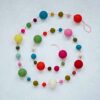 The Felt Small and Large Ball Garland, spiraled on a light surface, features vibrant red, pink, green, yellow, blue, and orange balls in various sizes for a lively decor touch.