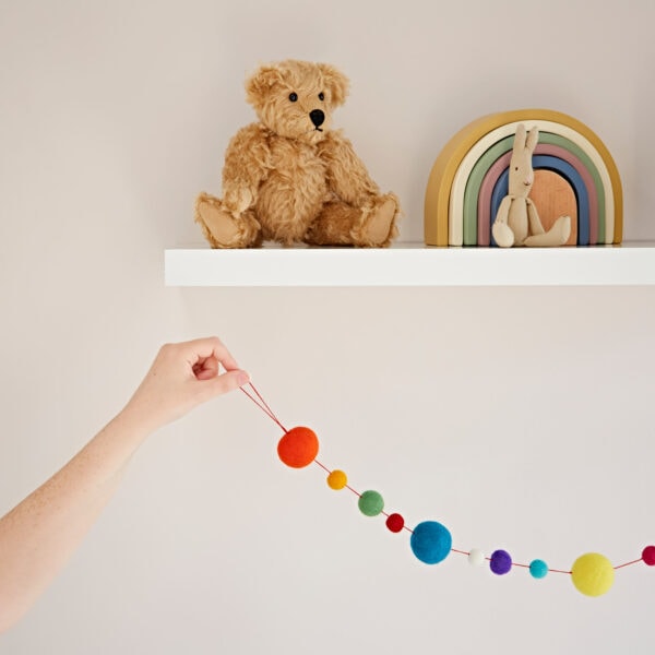 A hand holds a vibrant Felt Small and Large Ball Garland below a white shelf with a teddy bear, wooden rainbow toy, and small bunny figure.