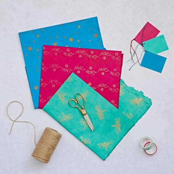 Colorful Lokta Paper sheets, scissors, gift tags with strings, a spool of twine, and rolls of tape are arranged on a light surface for perfect gift wrap.