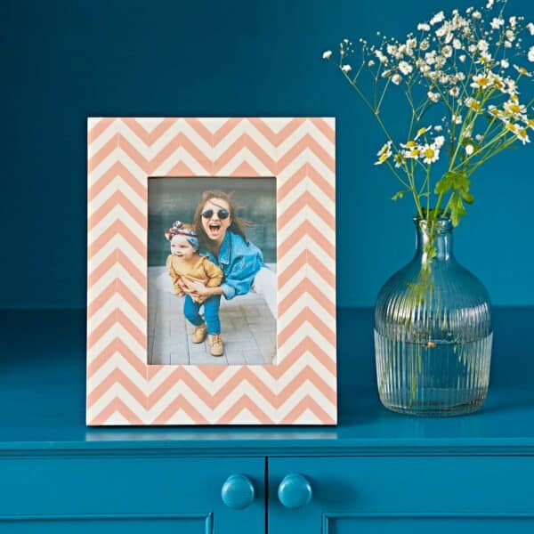 The Advika Pink Chevron Patterned Photo Frame with a picture of a woman and child stands on a blue cabinet beside a glass vase holding white flowers.