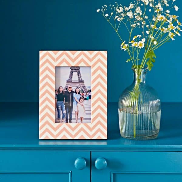 An Advika Pink Chevron Patterned Photo Frame with a family photo stands on a blue dresser beside a glass vase filled with white and yellow flowers.