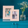 The Advika Pink Chevron Patterned Photo Frame and another chevron frame rest on a teal surface beside a clear ribbed vase with white flowers, set against a teal background.