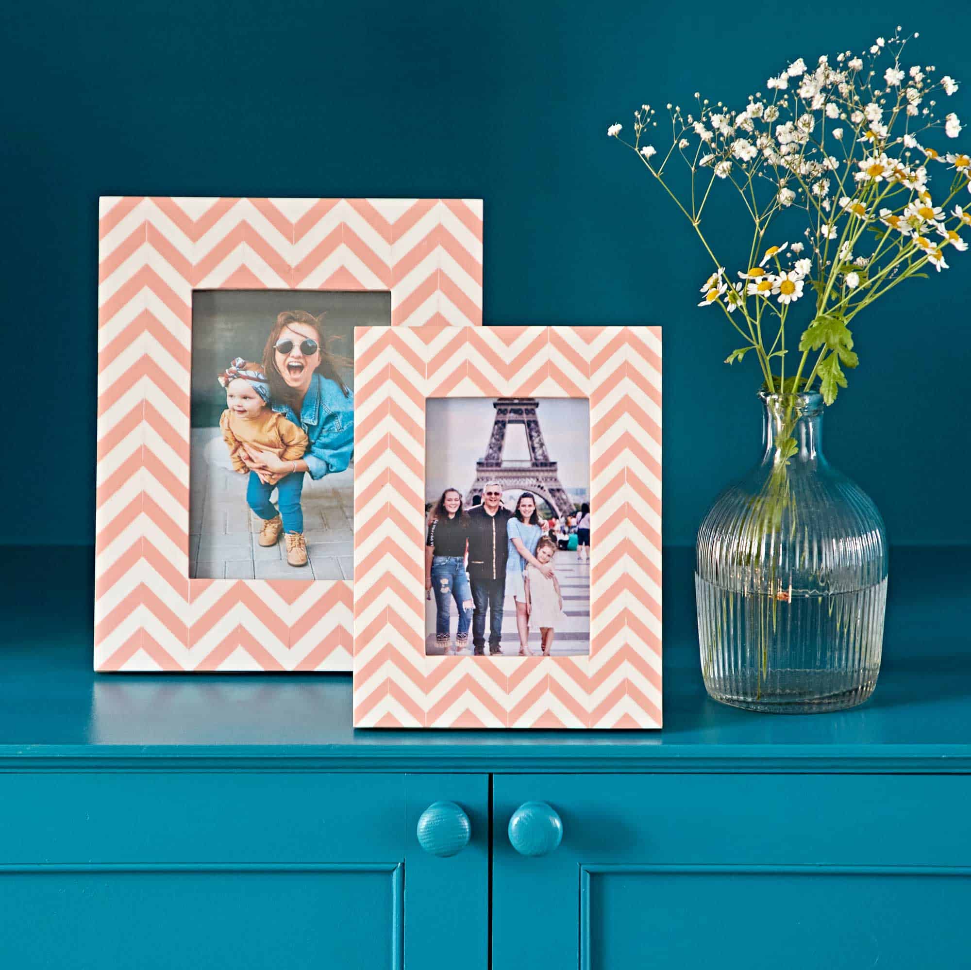 The Advika Pink Chevron Patterned Photo Frame and another chevron frame rest on a teal surface beside a clear ribbed vase with white flowers, set against a teal background.