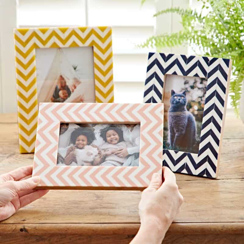Three chevron-patterned photo frames with family and pet photos are displayed on a wooden surface, with a plant and window in the background.