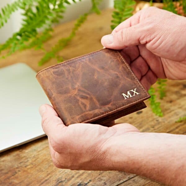 A person holds the Personalised Brown Distressed Buffalo Leather Billfold Wallet with "MX" initials embossed in the corner, beside a laptop and green plant on a wooden surface.