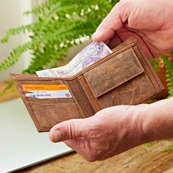 A person holds an open Personalised Brown Distressed Buffalo Leather Billfold Wallet containing British pound notes, bank cards, and a driver’s license, with a plant and laptop in the background.