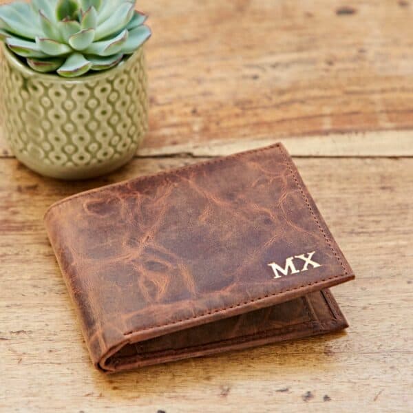 A Personalised Brown Distressed Buffalo Leather Billfold Wallet with gold "MX" initials rests on a wooden surface beside a small potted succulent.