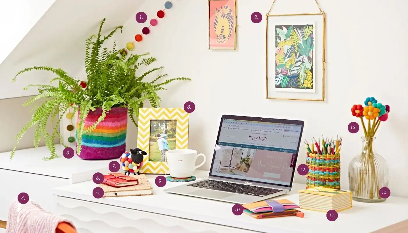A neat white desk with a laptop, colorful office supplies, a potted fern, framed photos, a mug, and decorative items under a slanted ceiling—The Ultimate Sustainable Home Office Setup for both style and productivity.