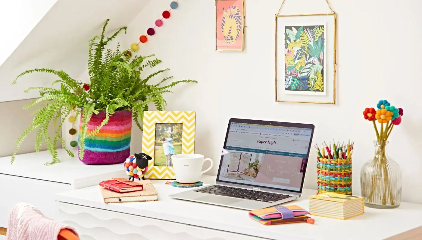 A colorful, decorated desk with a laptop, notebooks, pens, a mug, a framed photo, and a potted fern exemplifies The Ultimate Sustainable Home Office Setup under a slanting ceiling adorned with vibrant wall art.