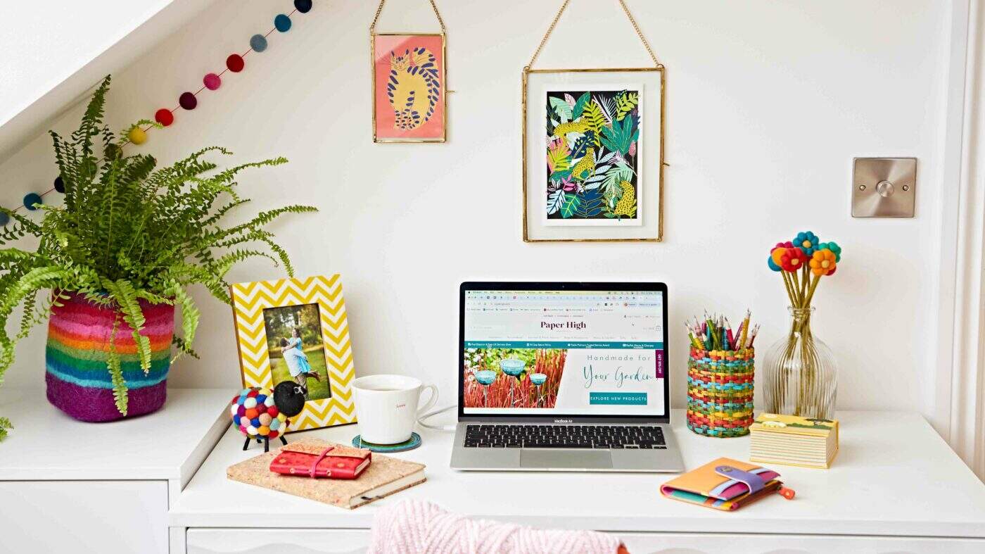 A neatly arranged desk under a sloped ceiling features a laptop, colorful stationery, framed art inspired by the Hampton Court Palace Garden Festival, a potted plant, and decorative items.