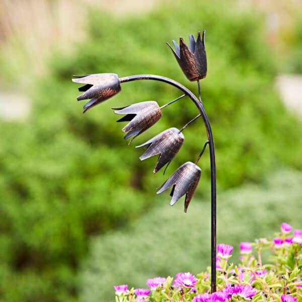 The Recycled Metal Echinacea Silhouette Garden Stake, featuring five bell-shaped flowers on a curved stem, stands among blooming pink flowers with green foliage in the background.